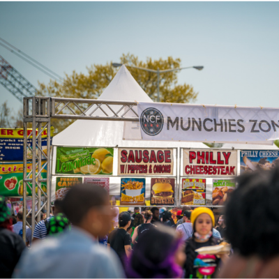 A bustling outdoor food festival scene featuring a “Munchies Zone” tent with signs for sausage, Philly cheesesteak, and lemonade, surrounded by a crowd of attendees.